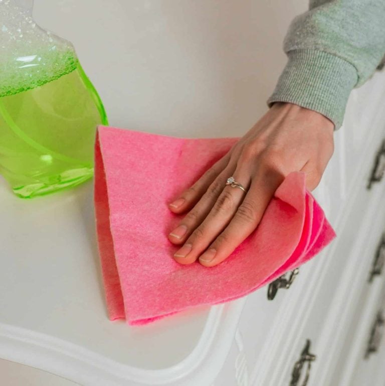 A woman's hand dusting a dresser with a pink cloth and a cleaning product spray bottle sitting on the dresser.